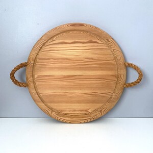 May include: A round wooden serving tray with rope handles. The tray is made of light wood with a natural finish.