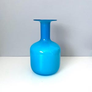May include: A vibrant, turquoise-colored glass vase. The vase has a rounded body and a long, slender neck that flares out at the top. The vase is approximately 10 inches tall and sits on a white surface against a gray background.