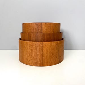 May include: A set of three cylindrical wooden bowls with a natural light brown wood grain finish. The bowls are stacked vertically, with the largest bowl at the base, the medium bowl in the middle, and the smallest bowl at the top, creating a tiered structure.