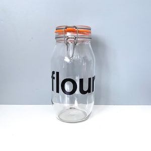 May include: A clear glass jar with a metal lid and an orange rubber seal. The jar has a black vinyl label that says "flour".