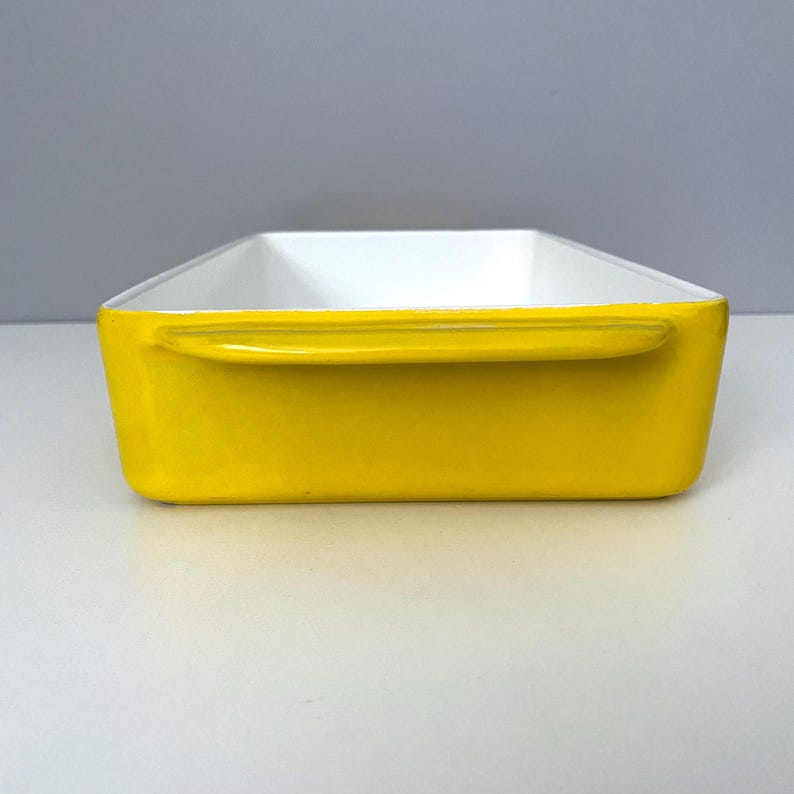 Op de afbeelding: Een rechthoekige ovenschaal met een heldergele buitenkant en een witte binnenkant. De schaal heeft aan elke kant een handvat. Het ontwerp is eenvoudig en modern, geschikt om in te bakken en te serveren.