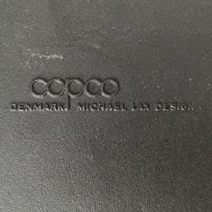 Op de afbeelding: Close-up van een getextureerd, donkergrijs oppervlak met het in reli&euml;f aangebrachte woord "copco" en de tekst "DENMARK MICHAEL LAX DESIGN". Het oppervlak lijkt deel uit te maken van een keukengerei-artikel.
