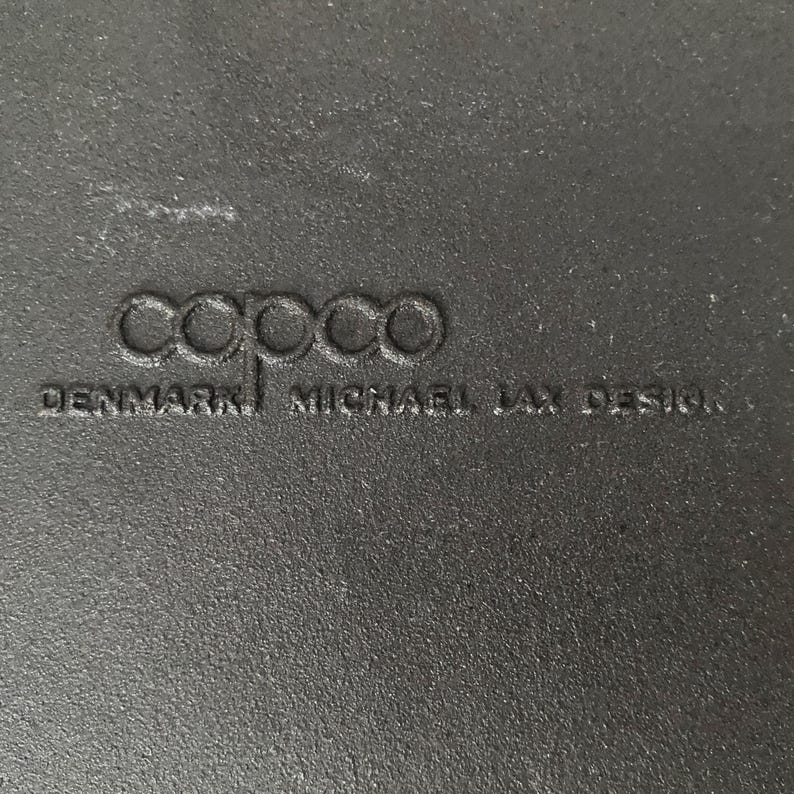 Op de afbeelding: Close-up van een getextureerd, donkergrijs oppervlak met het in reli&euml;f aangebrachte woord "copco" en de tekst "DENMARK MICHAEL LAX DESIGN". Het oppervlak lijkt deel uit te maken van een keukengerei-artikel.