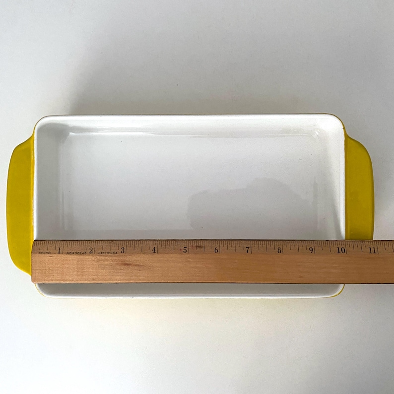 Op de afbeelding: Rechthoekige witte keramische ovenschaal met gele handvatten. De schaal is ongeveer 28 cm lang. De binnenkant is wit en de handvatten zijn felgeel. Een houten liniaal is naast de schaal geplaatst.