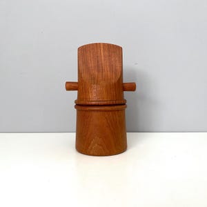 May include: A cylindrical wooden container with a warm brown grain, featuring a slightly curved top section and two small horizontal handles protruding from the sides. The object has a smooth finish and a distinct seam separating the top and bottom parts.