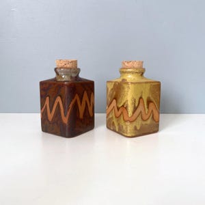 May include: Two ceramic jars with cork stoppers. The jars are square shaped and have a brown and yellow glaze with a wavy pattern.