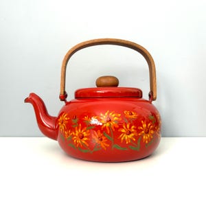 May include: A bright red enameled metal tea kettle featuring a hand-painted floral pattern of orange and yellow flowers with green stems. The kettle includes a curved spout, a removable lid with a wooden knob, and a tall, arched wooden handle.
