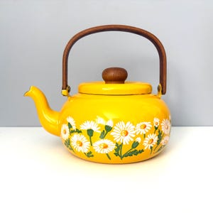 May include: A bright yellow enameled metal tea kettle featuring a white daisy floral pattern around the base. The kettle has a curved spout, a removable lid with a round wooden knob, and a tall, arched wooden handle attached with metal hardware.