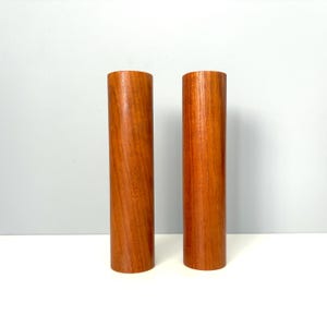 May include: Two cylindrical wooden objects with a warm, reddish-brown tone and a visible vertical wood grain pattern. Each piece features a smooth, straight-sided design with flat top and bottom edges, showcasing the natural texture of the wood.