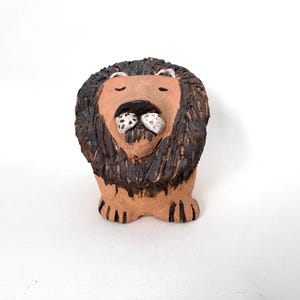 May include: A handcrafted ceramic lion figurine. The lion has a tan face and paws, a dark brown mane, and white and tan details on the mouth. The figurine is standing upright on a white surface.