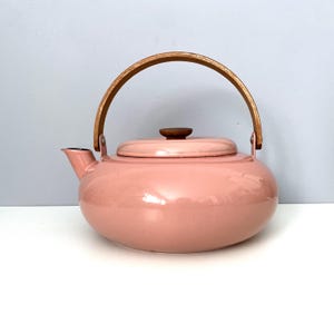 May include: A rounded, light pink teapot with a smooth, glossy finish. It features a curved wooden handle attached to the sides and a matching circular wooden knob on the center of the lid. The spout is short and angled upward on the left side.
