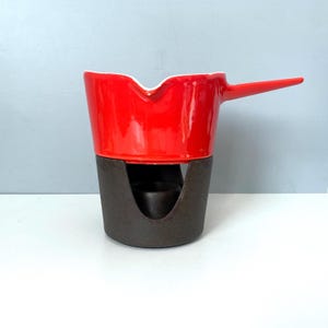 May include: A red ceramic fondue pot with a long handle and a dark brown base. The pot has a white interior and a glossy finish. The base has a circular opening for a heat source. A kitchenware item.