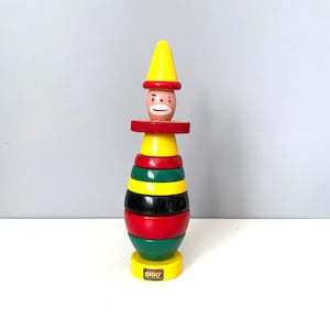May include: A vintage wooden stacking toy shaped like a clown with a yellow hat, red and black striped body, and a green and yellow base. The toy is made by BRIO.