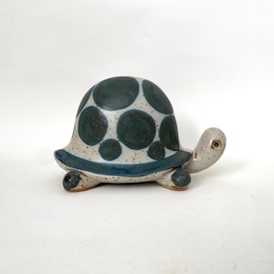 May include: A ceramic turtle figurine with a speckled white body, a dark teal shell with large, lighter teal spots, and a blue rim. The turtle has a small head and four stubby legs. The figurine is a decorative item.