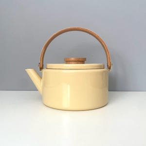 May include: A cream-colored enamel teapot with a wooden handle and lid knob. The handle is arched and made of light-colored wood, attached to the pot with metal brackets. The spout is angled, and the pot sits on a white surface.