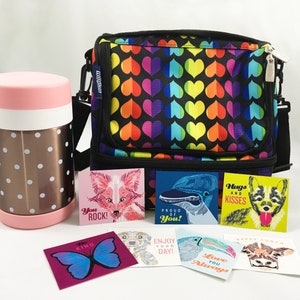 May include: A black lunch bag with a rainbow heart pattern. The bag has a zipper closure and a top handle. There are several small cards with encouraging messages and illustrations of animals, including a fox, a bird, a dog, and a cow.