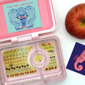 May include: A pink lunchbox with a clear compartment filled with colorful stickers of food items. The lunchbox is open and has a blue and green card with an elephant illustration and the text "YOU ARE AMAZING!" on top. A red apple and a blue card with a pink seahorse illustration and the text "I LOVE YOU!" are on the white surface.