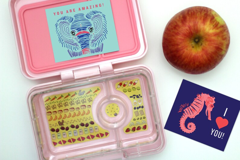 May include: A pink lunchbox with a clear compartment filled with colorful stickers of food items. The lunchbox is open and has a blue and green card with an elephant illustration and the text "YOU ARE AMAZING!" on top. A red apple and a blue card with a pink seahorse illustration and the text "I LOVE YOU!" are on the white surface.