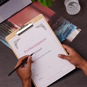 May include: A wooden clipboard with a silver clip holding a journal titled "Perimenopause Journal." The journal has a light pink border and text. A person is writing on the journal with a black pen. The journal is designed to track symptoms, sleep, mood & wellbeing.