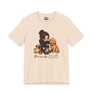 May include: A cream-coloured short-sleeve t-shirt featuring a graphic of a person feeding a pug, a kitten, and a golden retriever. The text "RD" is printed near the neckline, and the text "Proverbs 12:10" is printed in brown below the graphic.