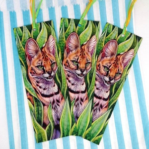 Puede incluir: Tres marcapáginas con una pintura de acuarela de un gato serval asomándose desde detrás de hojas verdes. El gato tiene un pelaje manchado y ojos verdes brillantes.