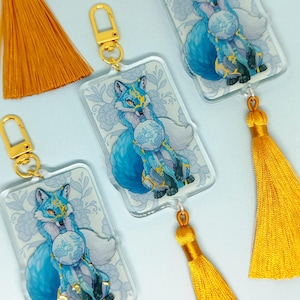 May include: Acrylic keychains featuring a blue fox design with gold accents and a white fluffy tail. The fox is holding a decorative orb. Each keychain has a gold-colored clasp and is paired with a bright orange tassel.