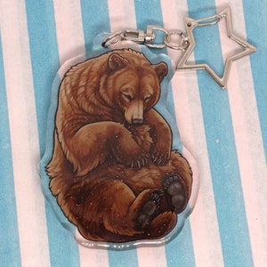 May include: A clear acrylic keyring featuring a brown bear curled up asleep. The bear has a glittery texture and is attached to a silver star-shaped clasp and a metal ring.