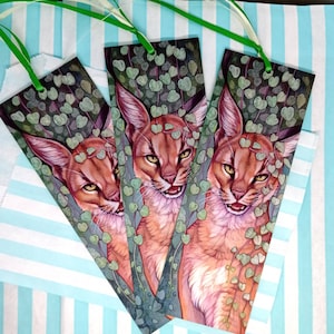 May include: Three illustrated bookmarks featuring a caracal cat with a green leafy vine design. Each bookmark has a green ribbon attached. The background is a light blue and white striped pattern.