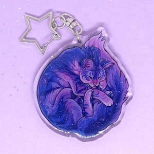 May include: A clear acrylic keyring featuring a sleeping cat with a galaxy design in shades of purple and blue. The cat is curled up, with a silver star-shaped charm and a clasp attached.