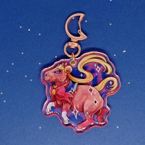 May include: A pink capybara dressed as Sailor Moon with a gold crown and a pink bow. The capybara is standing on a blue background with stars and a crescent moon. The capybara is a keychain charm.