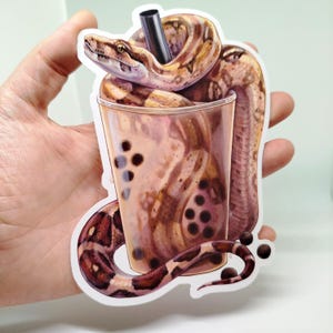 May include: A sticker of a brown and tan snake wrapped around a clear plastic cup filled with a brown liquid and black boba pearls. The snake has a black straw in its mouth.