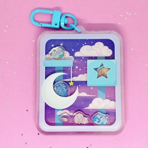 May include: A rectangular keyring with a dreamy night sky design. The keyring features a crescent moon, clouds, stars, and sleeping animal figures in blue, purple, and white. It has a light blue clasp.