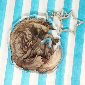 Puede incluir: Un llavero acrílico transparente con una ilustración de una hiena durmiendo. La hiena está representada en tonos marrones y beige con rayas. El llavero tiene un dije plateado en forma de estrella y un cierre plateado.