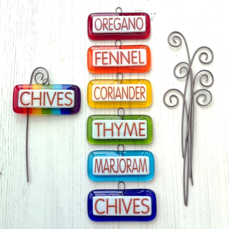 May include: A set of six colourful glass herb markers with metal hangers. The markers are labelled "Oregano", "Fennel", "Coriander", "Thyme", "Marjoram", and "Chives".