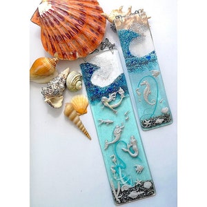 Può includere: Due ornamenti rettangolari in vetro con un tema oceanico blu e bianco. Un ornamento presenta una sirena e cavallucci marini, mentre l'altro presenta una sirena e pesci. Entrambi gli ornamenti hanno una finitura glitter argentata.