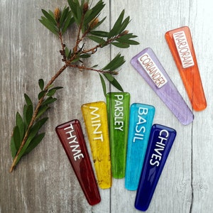 May include: A set of seven colourful glass plant markers with white lettering. The markers are labelled "Thyme", "Mint", "Parsley", "Basil", "Chives", "Coriander", and "Marjoram".
