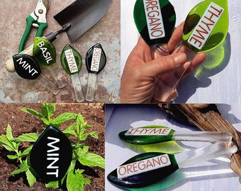 May include: Set of six glass plant markers in the shape of leaves. The markers are green and white with red lettering. The words on the markers are "Mint", "Basil", "Thyme", "Oregano", "Thyme", and "Oregano".