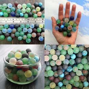 Peut inclure: Une collection de billes en verre colorées dans différentes nuances de bleu, vert et blanc. Les billes sont disposées dans un bol et sur une surface en bois. Certaines billes ont une finition givrée ou irisée.