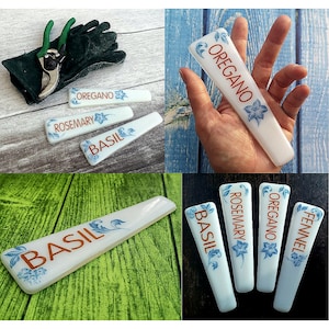 May include: Set of six white ceramic plant markers with blue floral designs and orange text. The markers are labelled "Basil", "Rosemary", "Oregano", "Fennel", "Basil", and "Oregano".