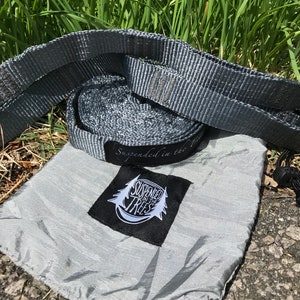 May include: A gray hammock suspension system with a matching carrying bag. The system includes a woven strap and a small bag with the text "Suspended in the Trees" and a logo. The items are displayed on a grassy surface.
