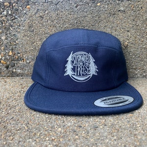 May include: Navy blue five-panel cap with a flat brim. The cap features white embroidered text and a tree design that reads "SUSPENDED IN THE TREES". The cap has an adjustable strap and a Yupoong tag.