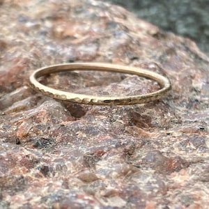Könnte beinhalten: Ein dünner, gehammerter Goldring liegt auf einer strukturierten, mehrfarbigen Felsfläche. Der Ring hat eine zarte, gehammerte Textur um das Band. Ein schlichtes, elegantes Schmuckstück.
