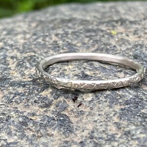 Könnte beinhalten: Ein silberner Ring mit einem strukturierten geometrischen Muster. Der Ring ist dünn und liegt auf einer grauen Steinoberfläche. Das Design des Rings weist kleine, sich wiederholende Formen um das Band auf.