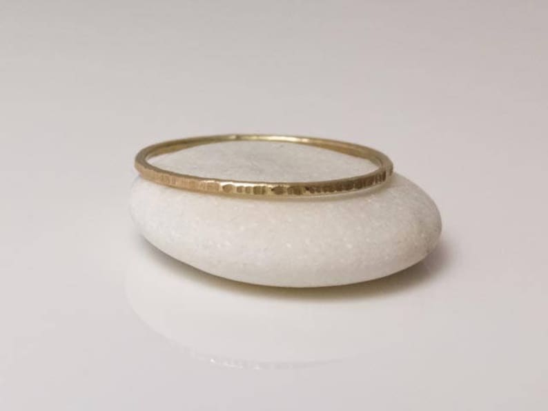 K&ouml;nnte beinhalten: Ein d&uuml;nner goldener Ring mit geh&auml;mmerter Textur, der auf einem wei&szlig;en Stein liegt.