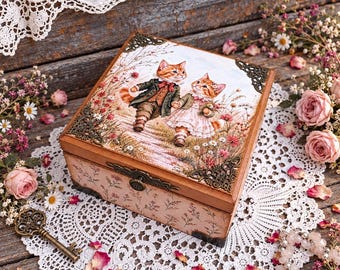 Puede incluir: Una caja de madera cuadrada con una ilustración de dos gatos antropomórficos con ropa formal caminando por un campo de flores. La caja tiene esquinas metálicas ornamentadas, un cierre de metal y un patrón floral rosa claro en los lados.