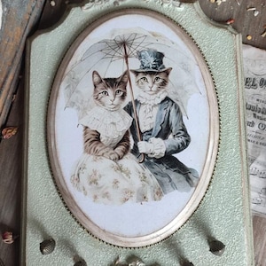May include: A vintage-style key holder with a green painted frame and ornate silver detailing. The frame features a circular image of two cats wearing hats and holding an umbrella. The key holder has four decorative metal knobs.