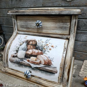 May include: Wooden bread box with a distressed finish. The front features a watercolor painting of bread, a milk bottle, and flowers. It has a drawer with a decorative knob and a curved, roll-top design.