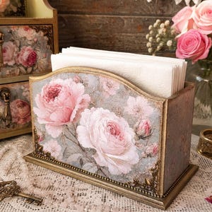 Puede incluir: Un servilletero rectangular de madera con un estampado decorativo de rosas rosas sobre un fondo gris. El soporte tiene bordes dorados con detalles metálicos ornamentados en las esquinas y contiene servilletas de papel blancas.