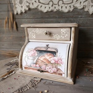May include: A wooden bread bin with a drawer and a hinged door. The box is painted in a light beige colour with floral stencilling. The door features a watercolour illustration of croissants, flowers, and a teapot.