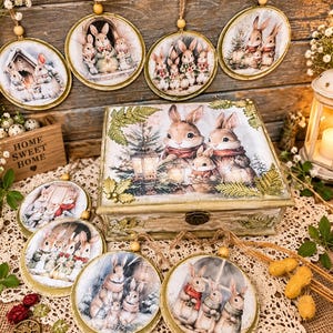 Pode incluir: Uma caixa retangular de madeira e vários ornamentos circulares com ilustrações de coelhos em cenários de inverno. A tampa da caixa exibe uma família de coelhos perto de uma lanterna e um pinheiro. Os ornamentos pendem de cordões com contas.